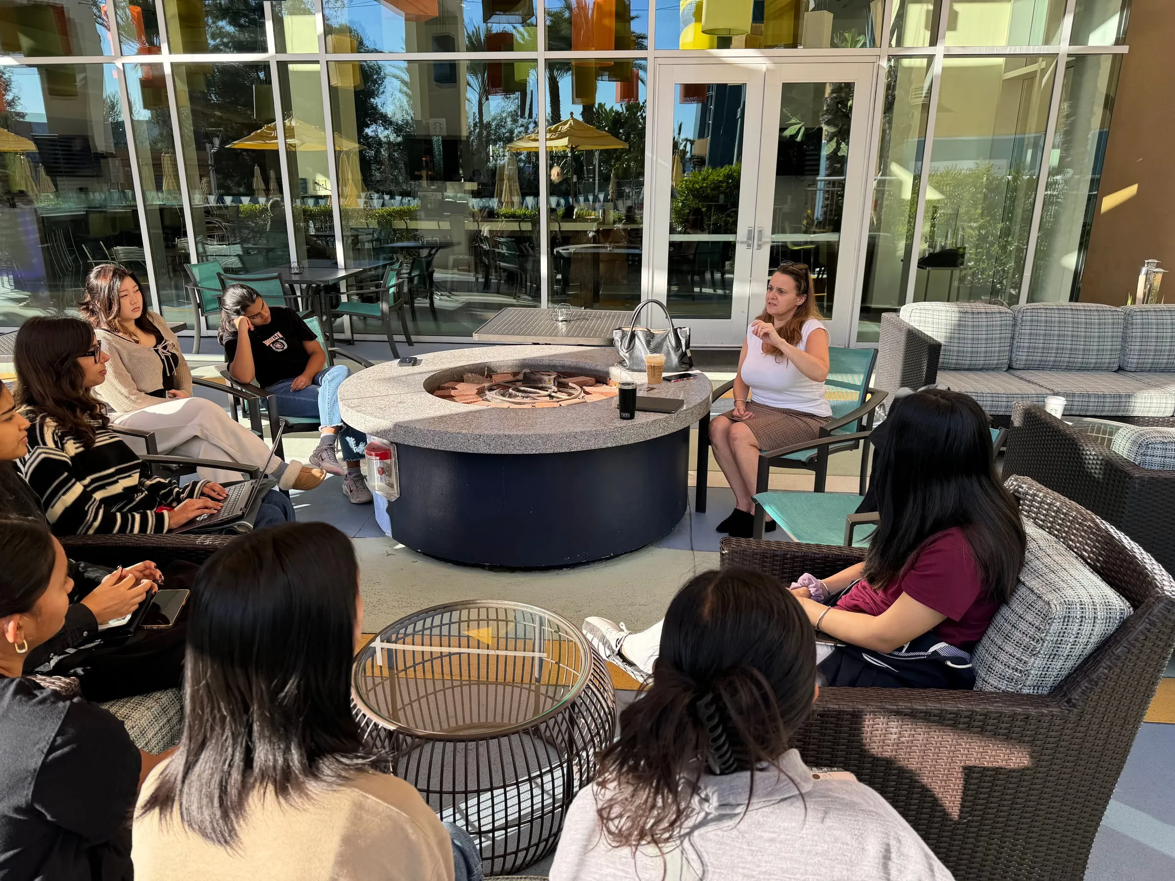 Members sit in patio chairs around a round stone fire pit outside a hotel while a woman opposite them speaks, one member taking notes on a laptop
