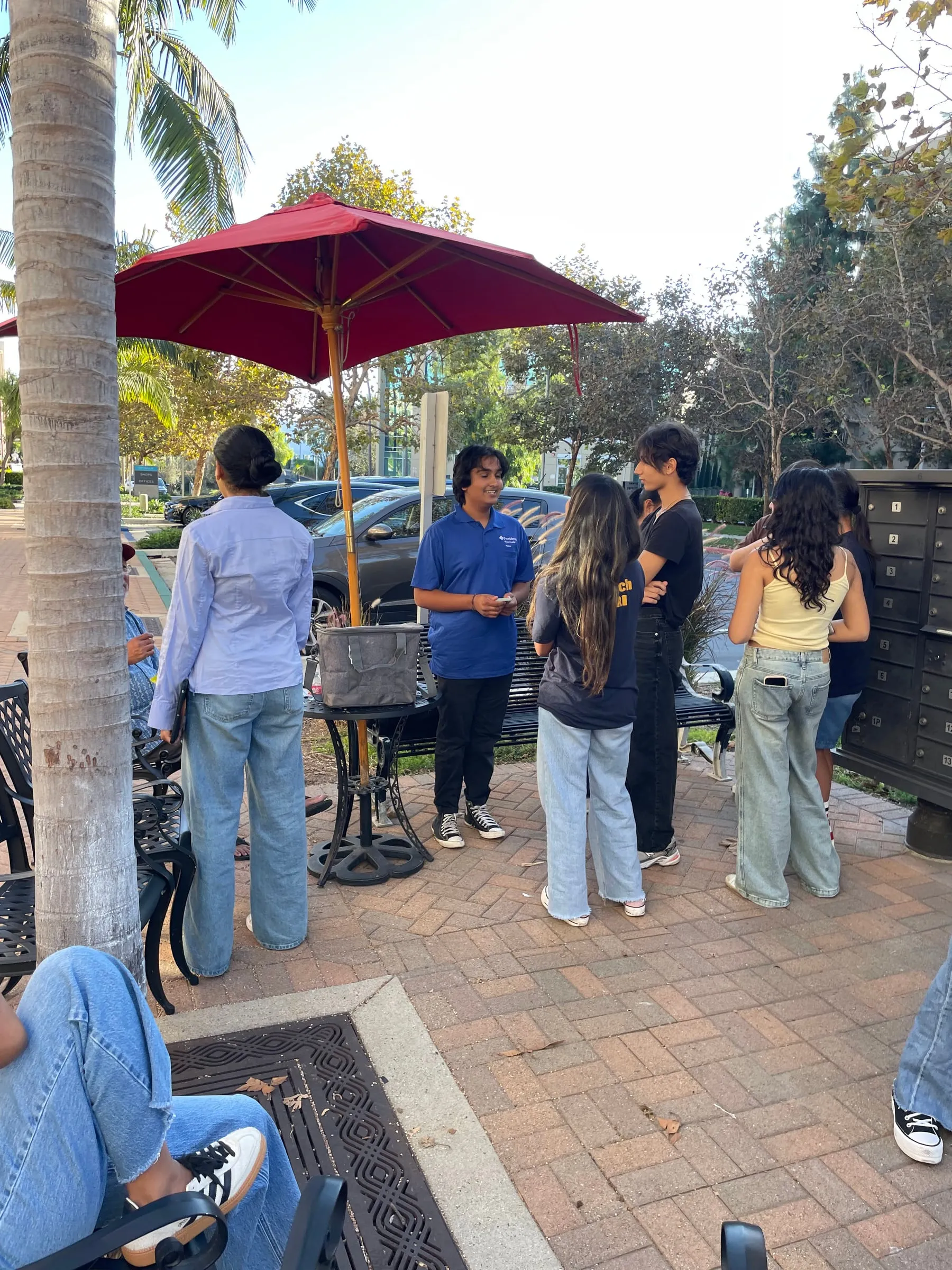 Teenagers stand talking together on a brick patio beside a red café umbrella, one of them in a blue volunteer polo shirt addressing the group