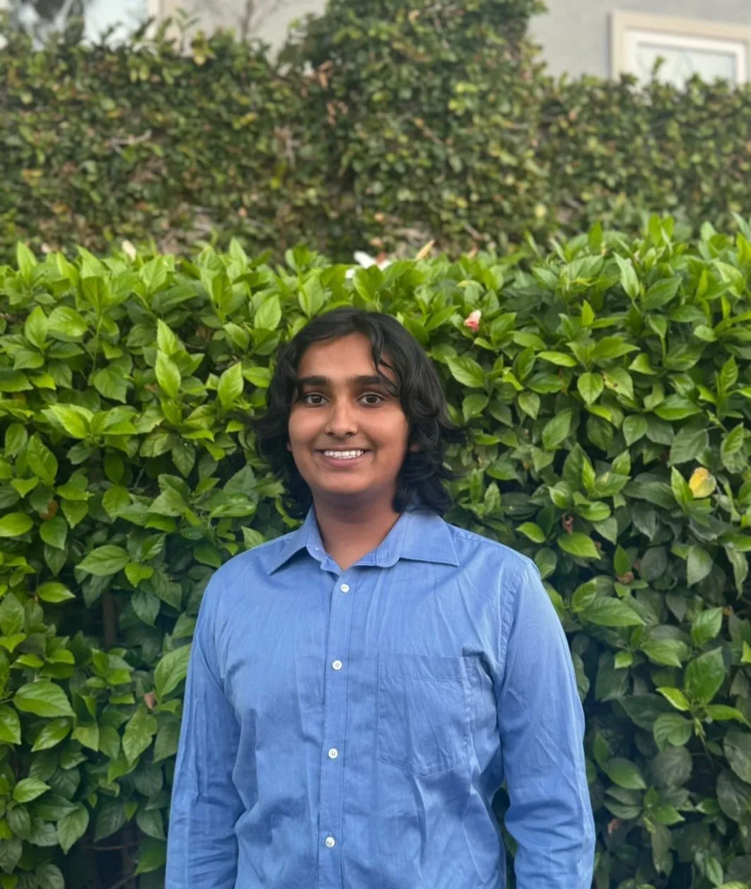 Ansh Sharma in front of a tall green hibiscus hedge, wearing a light blue button-down shirt