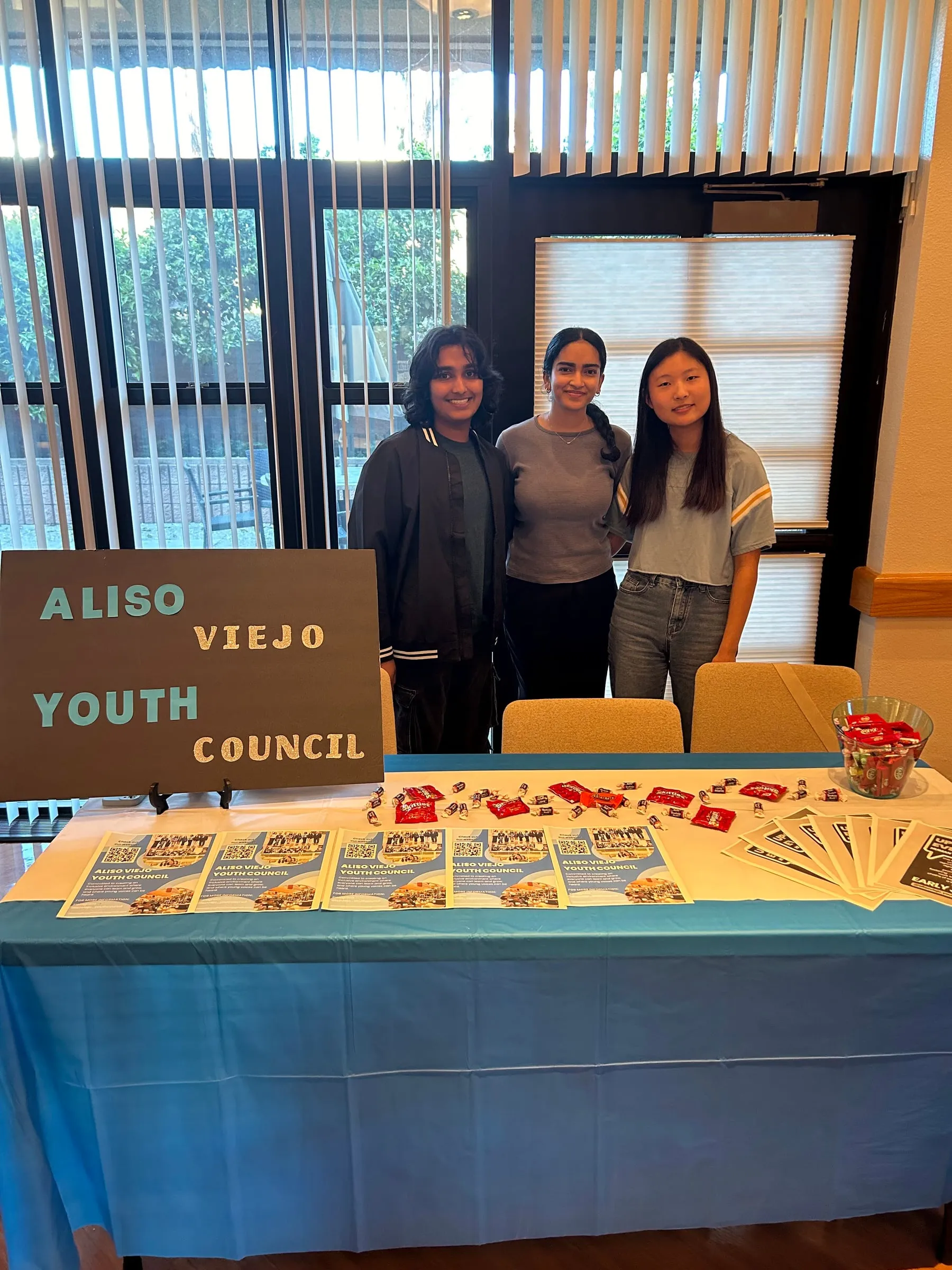 Three council members stand behind a blue-draped table laid out with council flyers and bowls of candy, beside a hand-lettered "Aliso Viejo Youth Council" sign