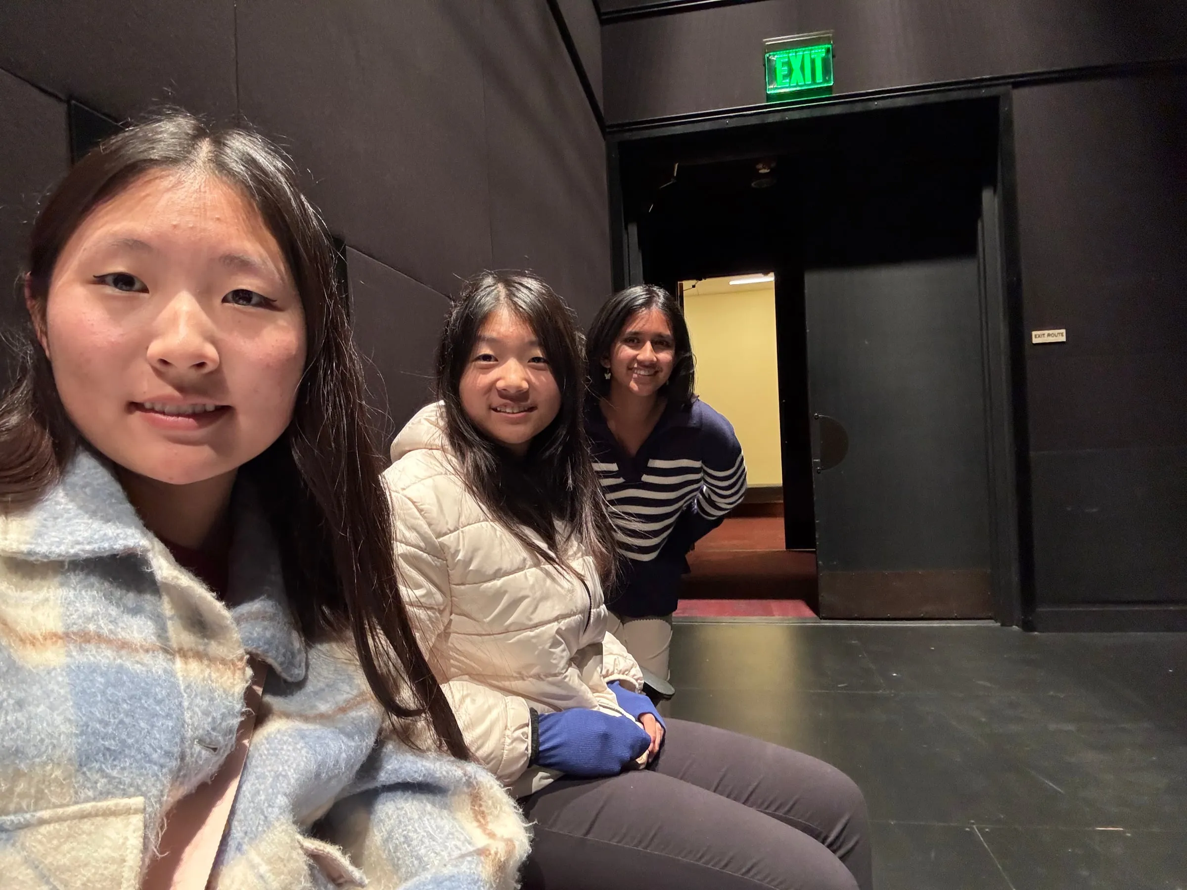Three council members sit together in the front row of a dark black box theatre, turning to smile at the camera beside a lit green exit sign