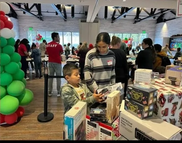 A teenage volunteer helps a young boy choose a boxed gift from a table of wrapped presents at a holiday event decorated with red and green balloon columns