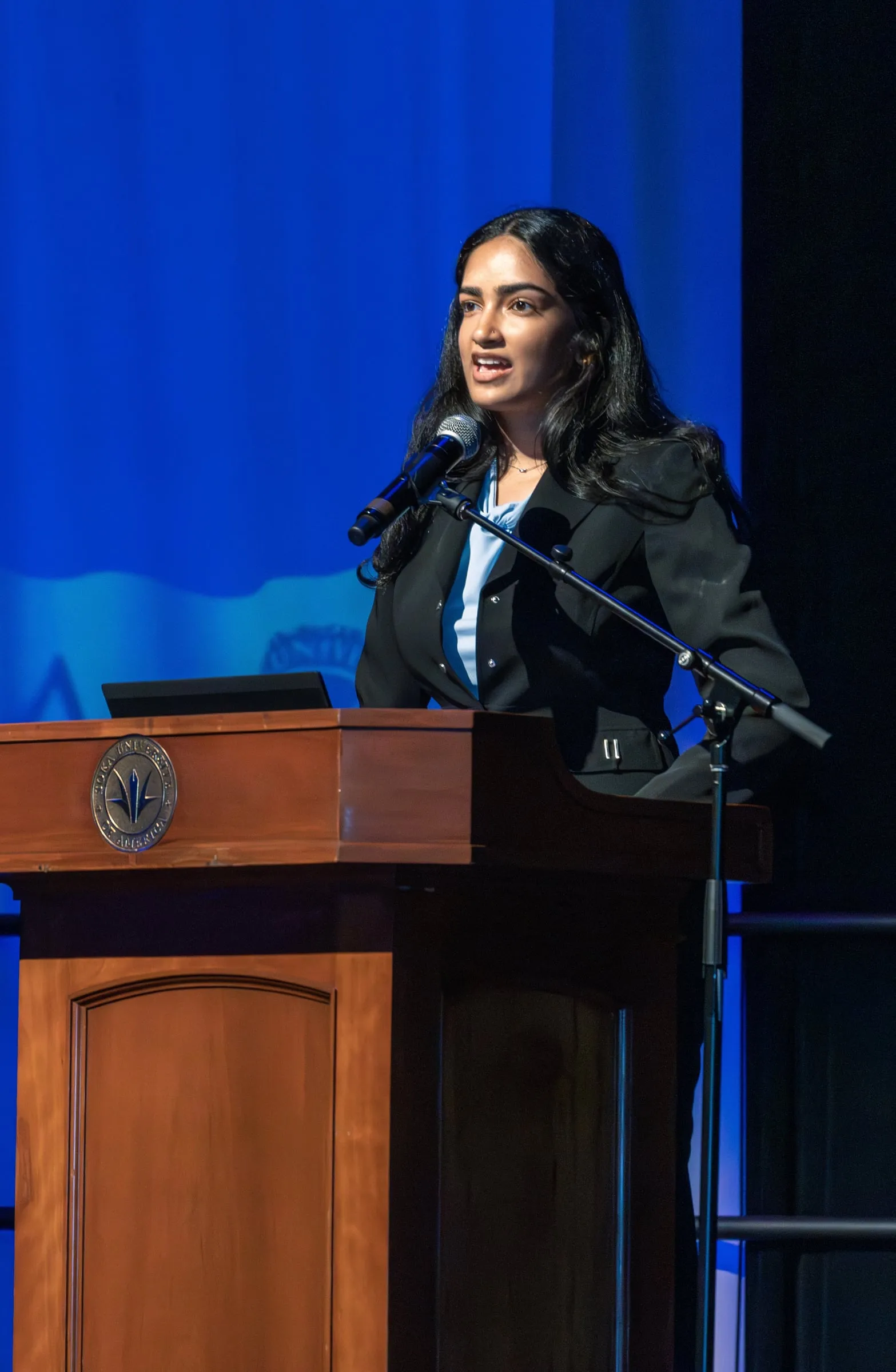 A council member in a black blazer speaks into a microphone from a wooden lectern bearing the Soka University seal, lit against a deep blue stage backdrop