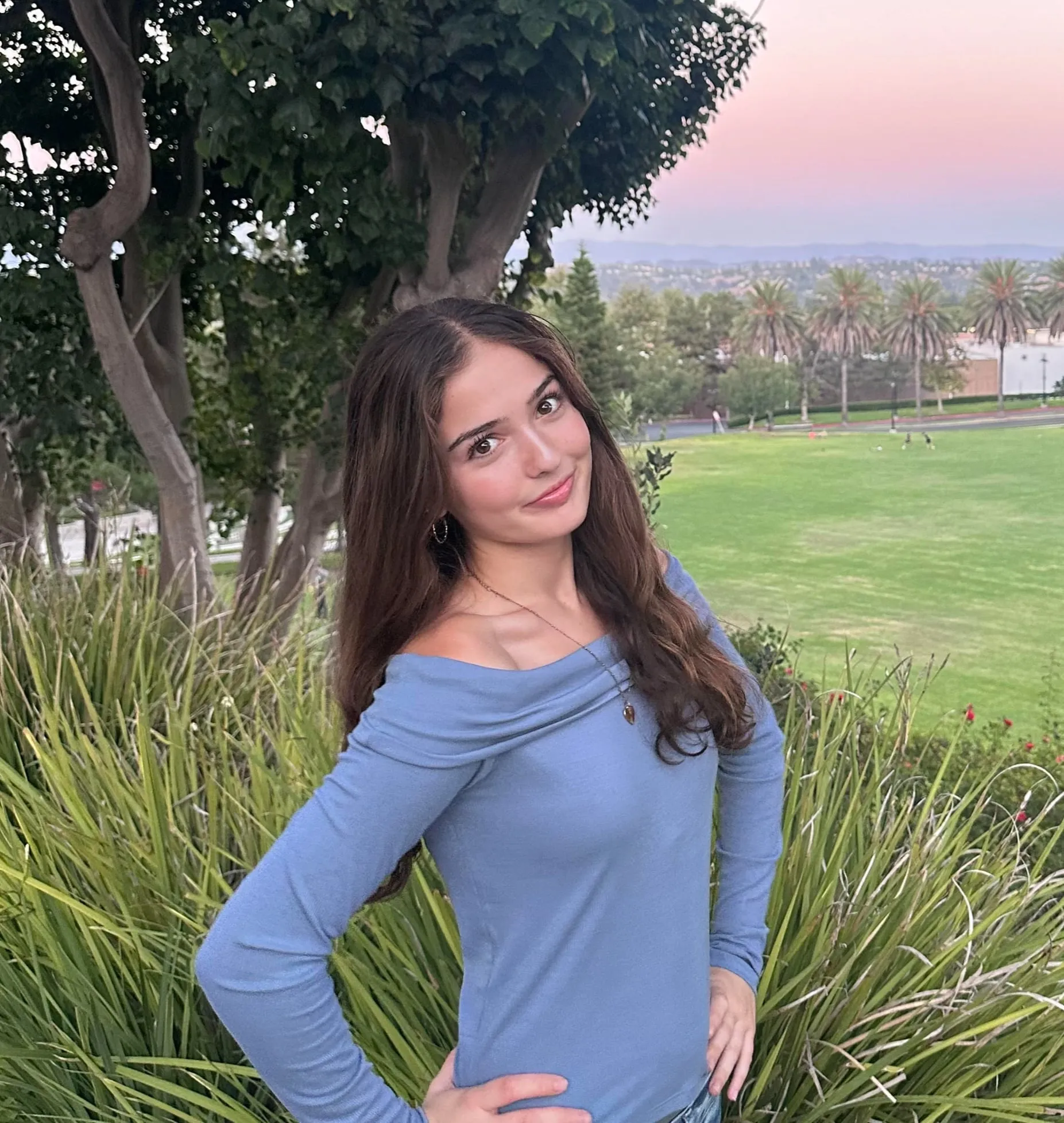 Kimia Ahmadi standing by tall grasses above a park lawn at dusk, wearing a blue off-the-shoulder top with one hand on her hip