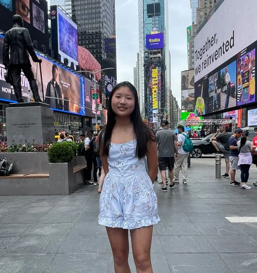 Olive Kim standing in Times Square among lit billboards, wearing a light blue floral sundress