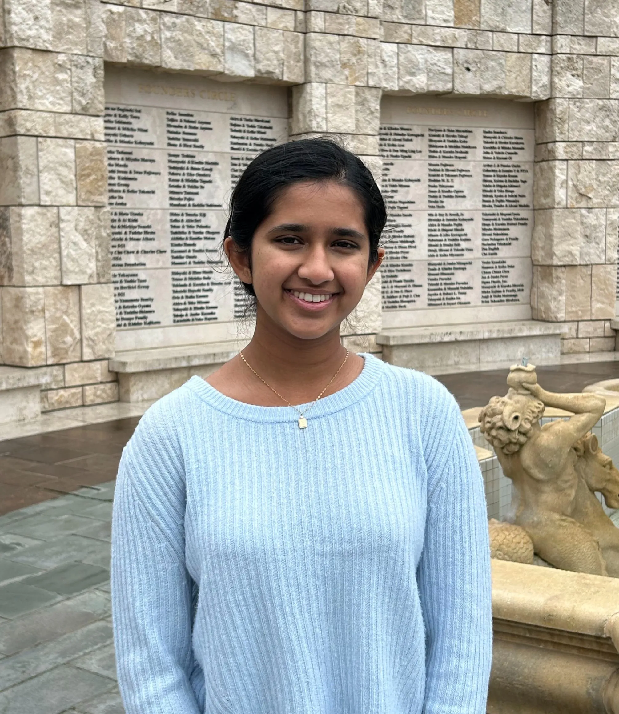 Carina Leo in a pale blue ribbed sweater, standing in a stone courtyard beside the Founders Circle wall and fountain
