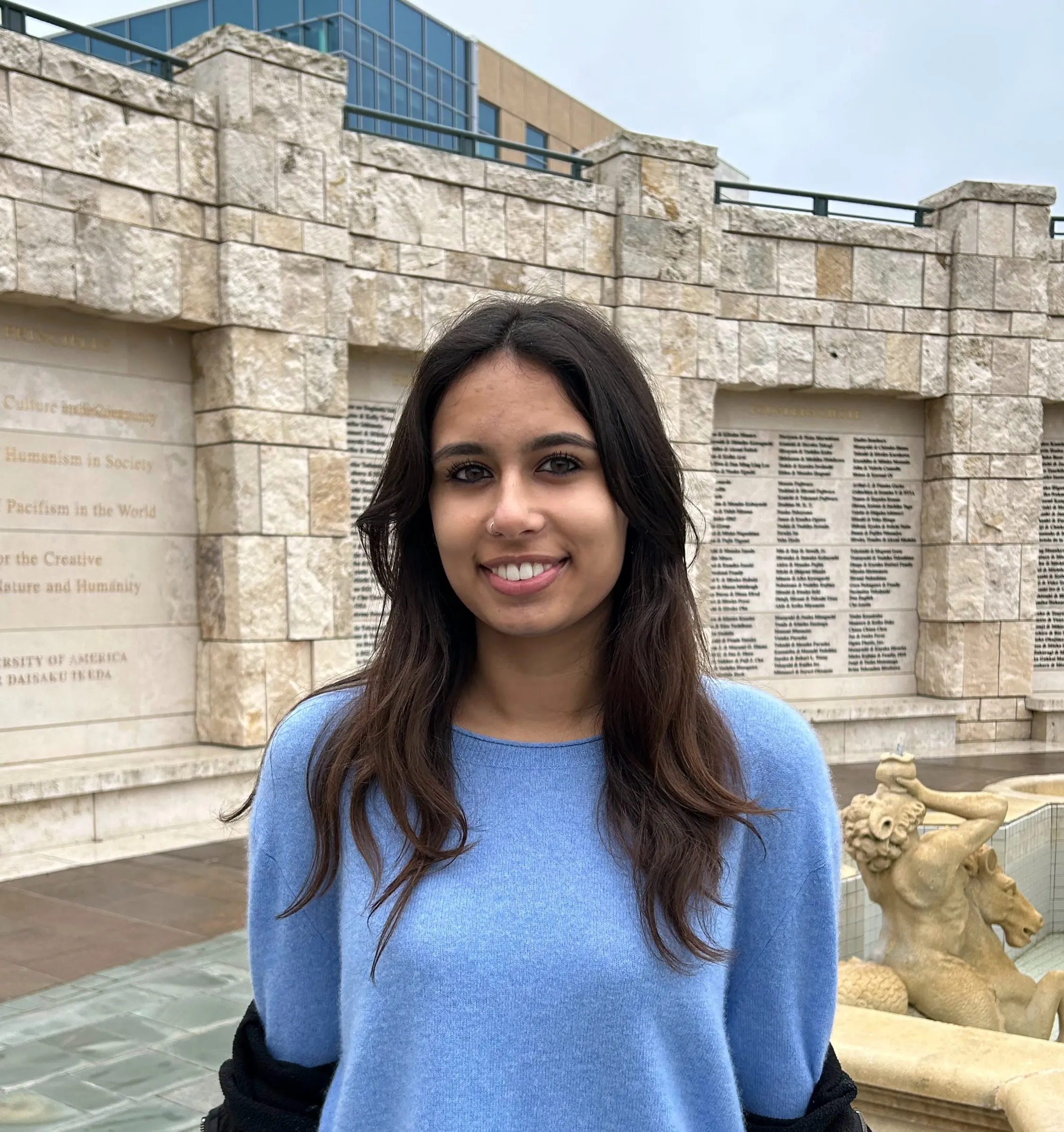 Maya D'lima in a light blue sweater, standing in a stone courtyard with an inscribed wall and fountain behind her