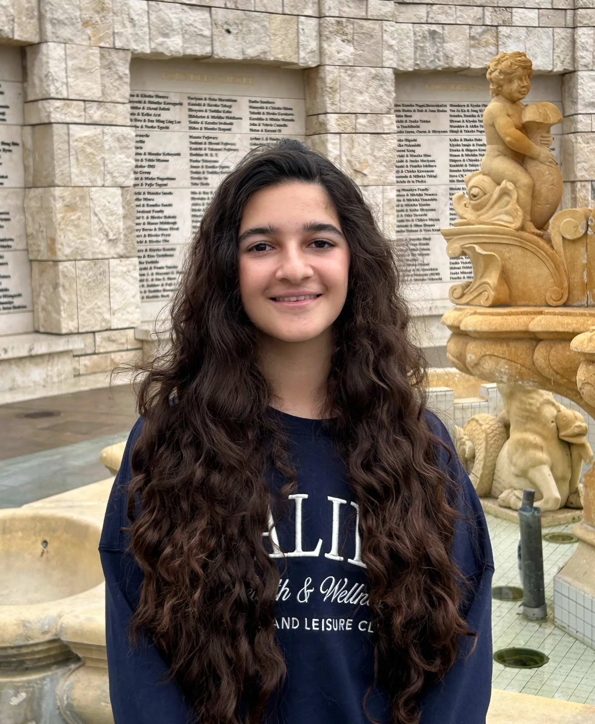 Rama Ketabchi in a navy embroidered sweatshirt, standing in a courtyard in front of a stone donor wall and fountain