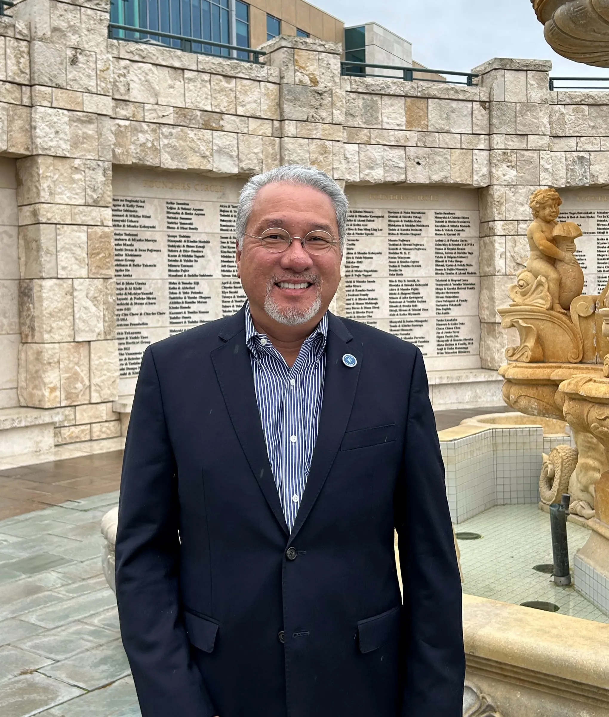 Ross Chun in a navy blazer and striped shirt, standing beside a stone Founders Circle donor wall and ornate fountain
