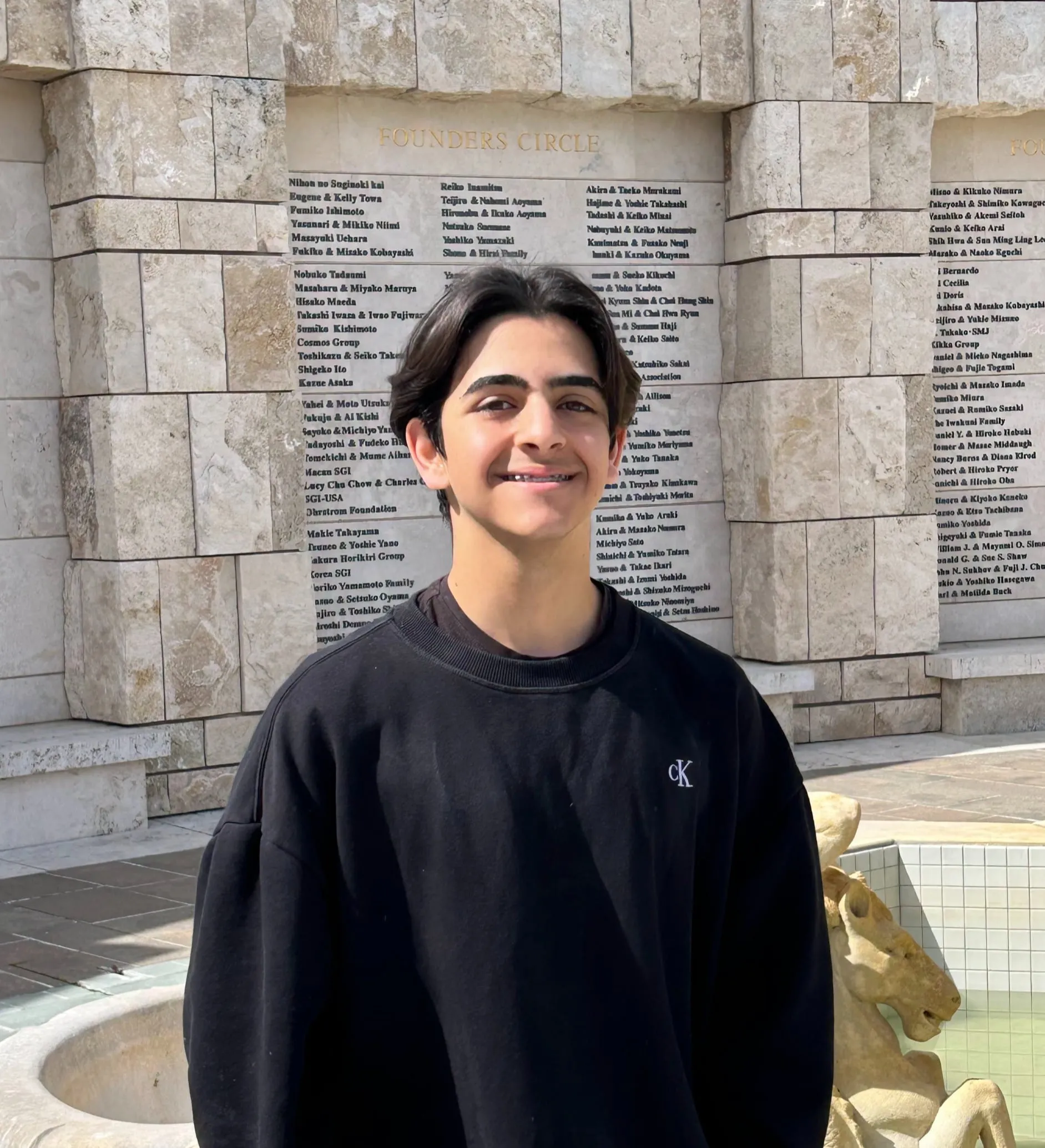 Ryan Ramlawi in a black crewneck sweatshirt, standing in front of the Founders Circle donor wall and fountain