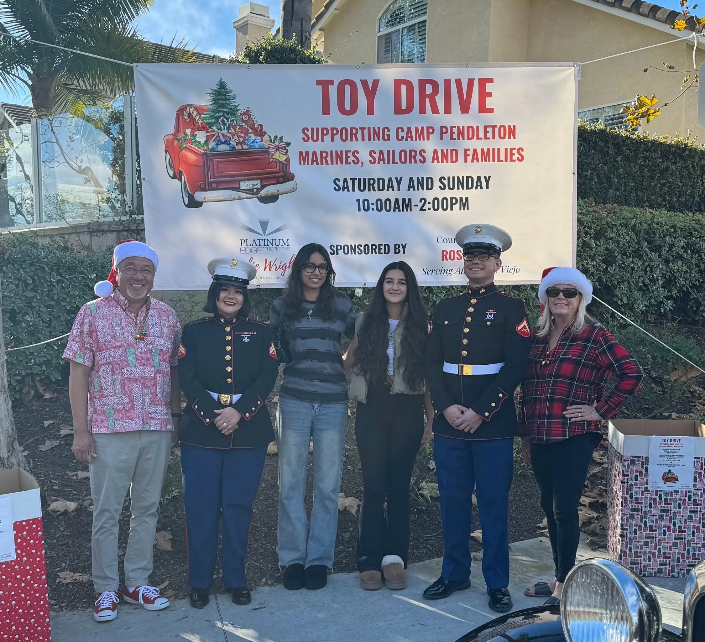 Six people stand in a driveway in front of a banner reading "Toy Drive — Supporting Camp Pendleton Marines, Sailors and Families", two of them in Marine dress uniform and two wearing Santa hats, with donation boxes at either side
